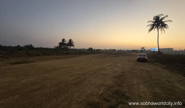 A wide-angle view of the construction site at Sobha World City in Hoskote, Bangalore, featuring cleared land, a parked car, and palm trees under a sunset sky.